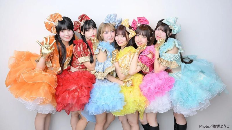 カラフルな衣装を着た7人組の女性アイドルグループの写真です。各メンバーは、オレンジ、赤、青、黄、ピンク、水色などの異なる色のドレスを着ています。全員がカメラに向かって微笑んでいます。