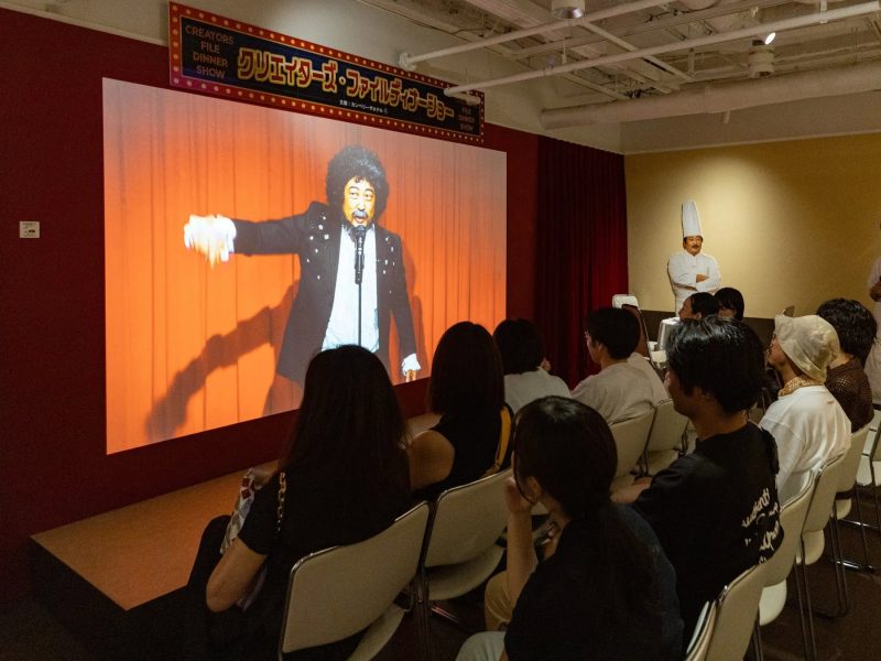 観客席で、スクリーンに映し出された人物の映像を見ている観客の様子。