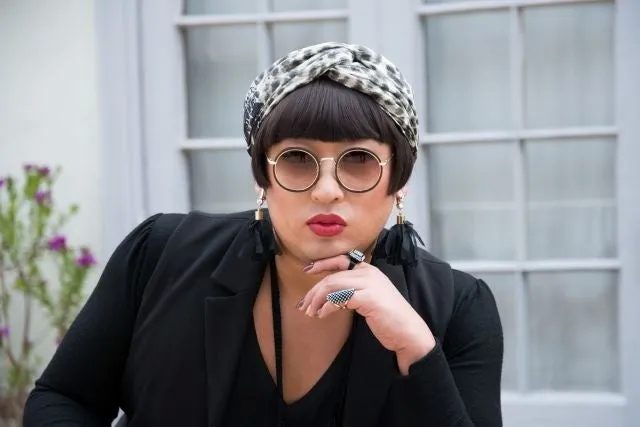 丸いサングラスをかけ、頭にスカーフを巻いた女性がカメラにポーズをとっている。