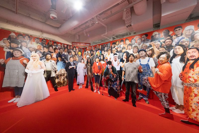 多くの人物が描かれた壁の前で、様々な格好をした人々が立っている写真。