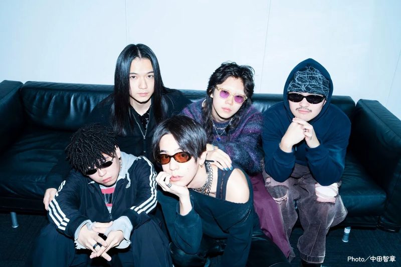 5人の男性が黒いソファに座ってポーズをとっている写真。彼らは皆、サングラスをかけており、さまざまなファッションスタイルをしています。