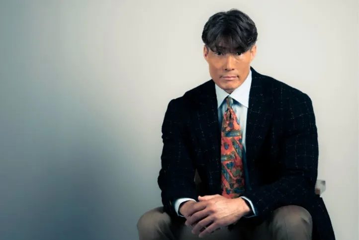 男性が椅子に座ってカメラ目線でポーズをとっている写真。彼はスーツを着ており、ネクタイをしています。