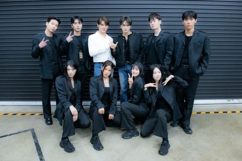 9人のグループがポーズをとって写真撮影をしている様子。一部のメンバーは黒いスーツを着ており、他のメンバーは白いシャツを着ている。