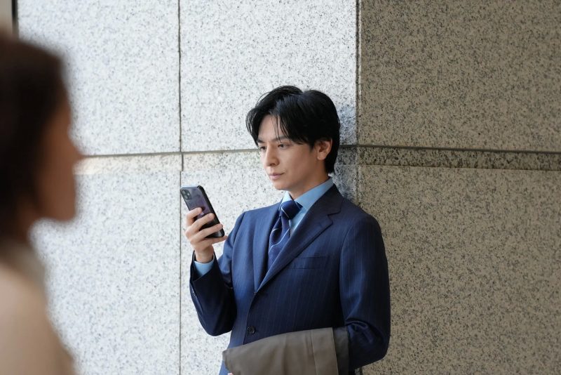 スマホを手に持つスーツ姿の男性が、壁に寄りかかっている様子。
