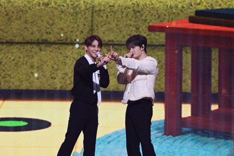 2人の男性がステージ上で歌を歌っている様子。2人ともマイクを持っており、笑顔で観客に手を振っている。背景には、赤と黄色の装飾が施されたステージセットがある。