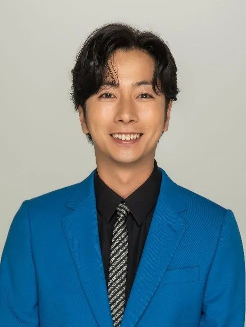 明るいグレーの背景の前で、青いジャケットと黒いシャツ、ストライプのネクタイを着用したアジア人男性が満面の笑みを浮かべているポートレート写真です。