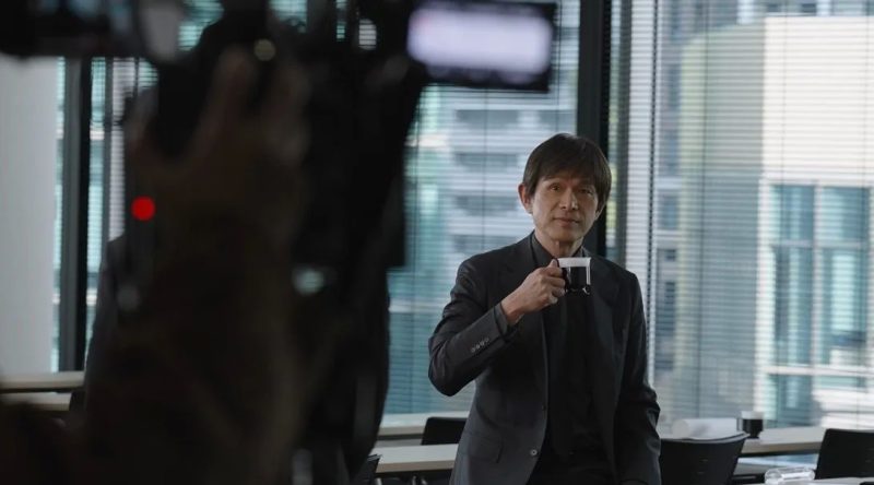 コーヒーカップを手にした男性がカメラの前で立っている写真です。彼は黒いスーツを着ており、窓のそばに立っています。