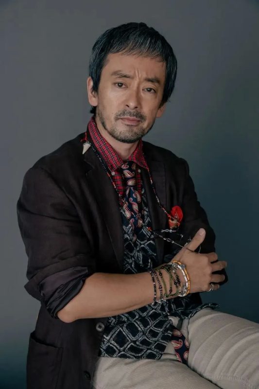 男性が椅子に座って腕を組んでいるポートレート写真。彼は茶色のジャケット、チェックのシャツ、ネクタイ、ブレスレットを着用しています。彼の顔にはひげとわずかに笑った表情です。