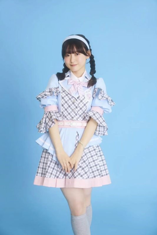 水色の衣装を着た女性のポートレート。彼女は、チェック柄のミニスカート、ピンクのリボン、髪に白いヘッドバンド、そして三つ編みをしている。彼女はカメラを見て微笑んでいる。