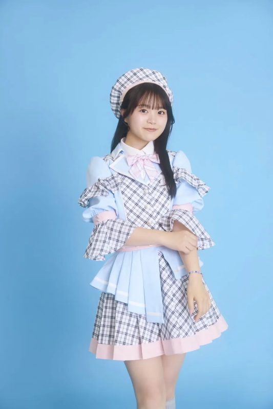 水色の背景に、チェック柄の衣装を着た若い女性が立っています。彼女はベレー帽をかぶり、腕を組んでカメラを見ています。