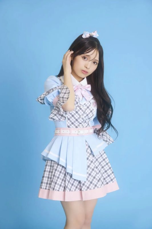 水色の背景に、ピンクのリボンとチェック柄の衣装を着た女性が立っています。彼女は髪を触り、カメラを見ています。