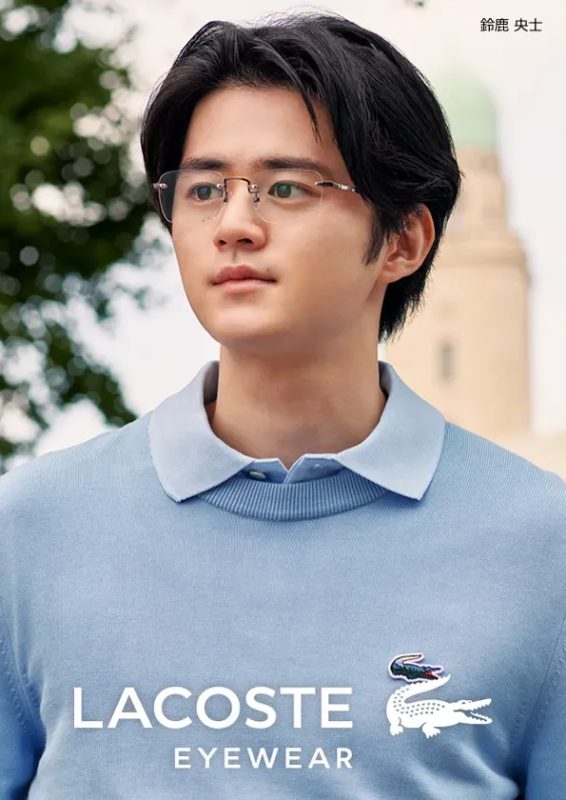 眼鏡をかけた男性が、水色のラコステのセーターを着て、カメラのほうを見ている写真